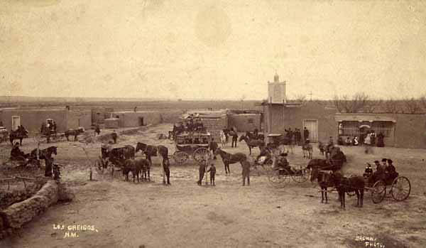 Old photo of New Mexico village, showing a stagecoach, wagons and carriages, and residents in Victorian clothing in front of adobe church and buildings.